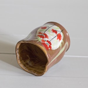Puede incluir: Una taza de cerámica marrón con un diseño pintado a mano de flores rojas y hojas verdes. La taza está esmaltada y tiene una forma ligeramente irregular.