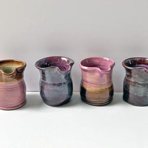 Könnte beinhalten: Vier kleine Keramikkrüge in einer Reihe angeordnet. Jeder Krug hat eine einzigartige Glasur in den Farben Rosa, Violett, Braun und Dunkelgrau. Die Gefäße haben einen bauchigen Körper, einen breiten Rand und einen kleinen integrierten Ausguss.