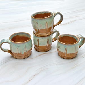 Può includere: Quattro tazze in ceramica fatte a mano con una glassa verde chiaro e marrone. Le tazze hanno una forma arrotondata e un manico.