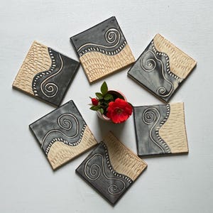 May include: Six square ceramic coasters arranged in a circle around a small red flower. Each coaster features a split design with a dark gray section containing a white spiral and a light beige section with a textured, carved pattern.