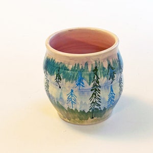 May include: A small, round ceramic mug with a light pink interior and a white exterior. The mug is decorated with a hand-painted design of a forest scene with blue and green trees.