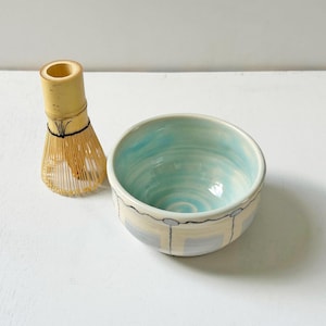 May include: A bamboo matcha whisk and a ceramic matcha bowl. The bowl has a light blue interior and a cream-colored exterior with gray and yellow geometric designs. The whisk is made of thin bamboo strips.