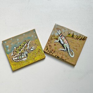 May include: Two square ceramic coasters with hand-painted animal designs. One coaster features a green and white frog with black spots on a yellow background. The other coaster has a lizard with red and white stripes.