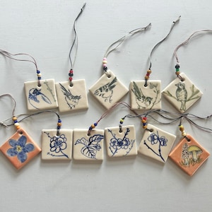 May include: A collection of ceramic ornaments with hand-painted designs. The ornaments feature birds, flowers, and mushrooms in blue and orange hues. Each square tile is strung with a cord and beads for hanging.