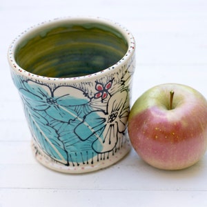 Puede incluir: Una taza de cerámica blanca con un diseño pintado a mano de hojas y flores azules y verdes. La taza está sentada junto a una manzana roja sobre una superficie blanca.