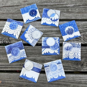 May include: A set of square ceramic tiles with blue and white illustrations. Each tile showcases a different design, including celestial bodies, waves, and landscapes. The artwork is hand-drawn, with a focus on line work and a limited colour palette.
