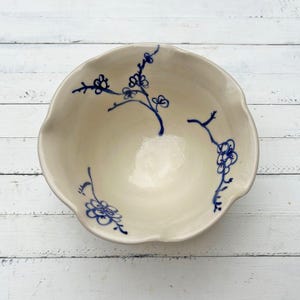 May include: Off-white ceramic bowl with a scalloped rim. The bowl is decorated with blue floral branch designs. The bowl is sitting on a white wooden surface.