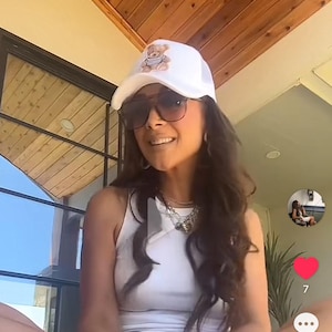 May include: A white trucker hat with a teddy bear graphic is worn by a person. The person is wearing sunglasses and a white tank top. A silver necklace is visible. The background includes a wooden ceiling and a window.
