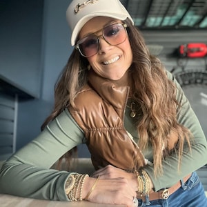 May include: A cream-colored baseball cap with a gold-colored horse bit design. The person is wearing brown-tinted sunglasses, a brown vest, and a green long-sleeved shirt. Multiple gold bracelets and a gold necklace are also visible.