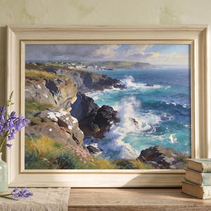 May include: A framed coastal landscape painting showing crashing waves and cliffs. The artwork is in a cream frame. A vase of bluebell flowers and books are on a wooden surface. The scene is reminiscent of the Cornish coast.