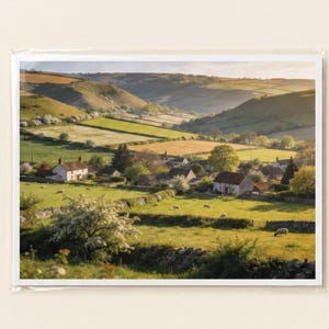 May include: A scenic landscape photograph of a rural village nestled in a valley. The image features rolling green hills, fields, and several houses with white walls and brown roofs. The scene is bathed in warm sunlight.