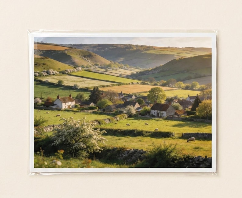 May include: A scenic landscape photograph of a rural village nestled in a valley. The image features rolling green hills, fields, and several houses with white walls and brown roofs. The scene is bathed in warm sunlight.