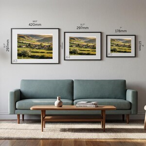 May include: Three framed landscape art prints of varying sizes (16.5" x 11.7", 11.7" x 11.7", and 7" x 5") hang above a teal sofa. The prints depict a rural scene with green fields and a village. A wooden coffee table and chair complete the room.