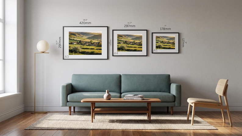 May include: Three framed landscape art prints of varying sizes (16.5" x 11.7", 11.7" x 11.7", and 7" x 5") hang above a teal sofa. The prints depict a rural scene with green fields and a village. A wooden coffee table and chair complete the room.