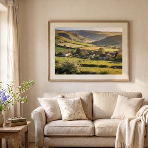 May include: A framed landscape print depicts a serene valley scene with rolling green hills, a village, and a bright sky. The artwork hangs above a beige sofa with throw pillows and a blanket. A wooden side table holds a vase of flowers.