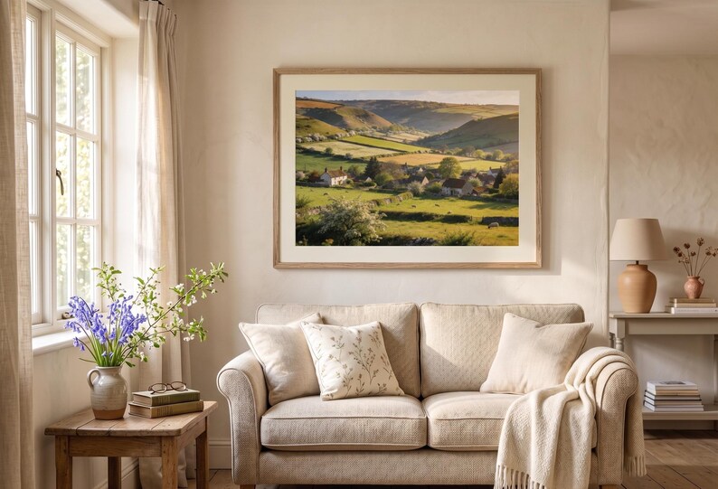 May include: A framed landscape print depicts a serene valley scene with rolling green hills, a village, and a bright sky. The artwork hangs above a beige sofa with throw pillows and a blanket. A wooden side table holds a vase of flowers.