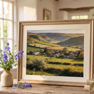 May include: A framed landscape print of a valley with rolling green hills, fields, and small houses. The frame is light brown. A vase of bluebell flowers sits on a wooden table in front of the print.