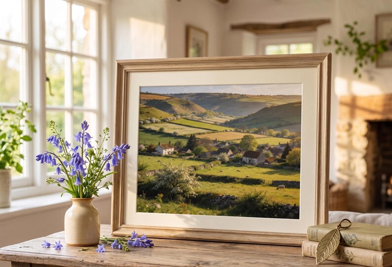 May include: A framed landscape print of a valley with rolling green hills, fields, and small houses. The frame is light brown. A vase of bluebell flowers sits on a wooden table in front of the print.