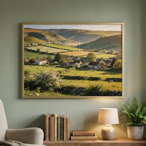 May include: A framed landscape print depicts a rural scene with rolling green hills, fields, and a small village. The artwork is in a gold frame and hangs on a sage green wall. A lamp, books, and a potted fern sit below the artwork.