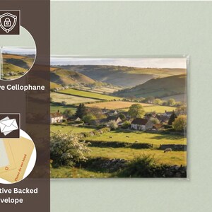 May include: A landscape print of a rural village scene, protected by cellophane and a protective backed envelope. The image shows rolling green hills, fields, and houses under a bright sky. The print is packaged for protection.