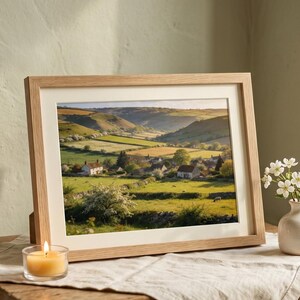 May include: A framed landscape print featuring a rural scene with rolling green hills, fields, and a village. The print is in a light wood frame, displayed on a wooden table with a lit candle and a small vase of flowers. A potted plant sits on a windowsill.