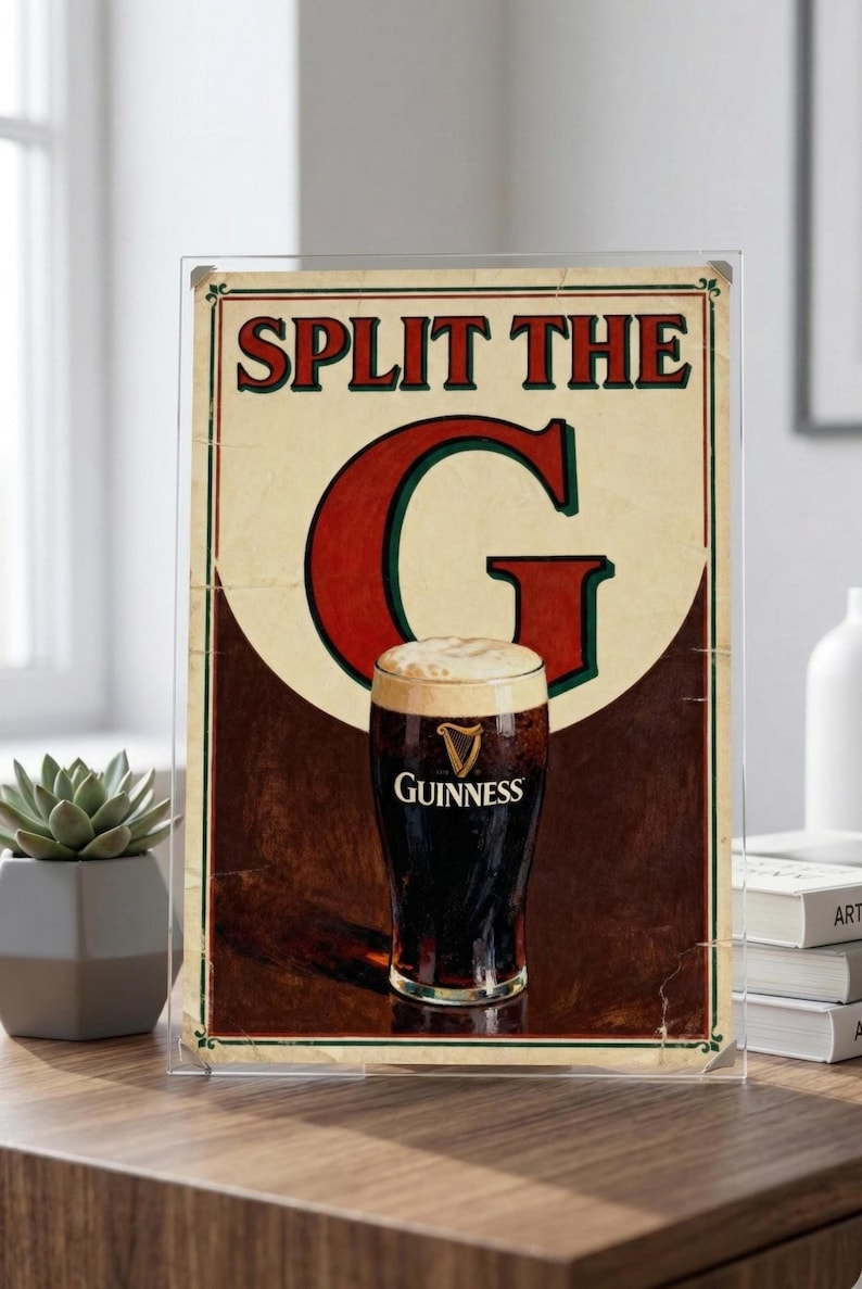 May include: Framed vintage-style Guinness beer advertisement. The sign reads "Split the G" above a pint of dark stout with a creamy head. The Guinness logo is visible on the glass. The background is a mix of cream, brown, and red.