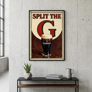 May include: Framed vintage-style Guinness beer advertisement. The poster features a pint of Guinness with a creamy head, and the text "SPLIT THE G". The poster is framed in black and hangs on a concrete wall.