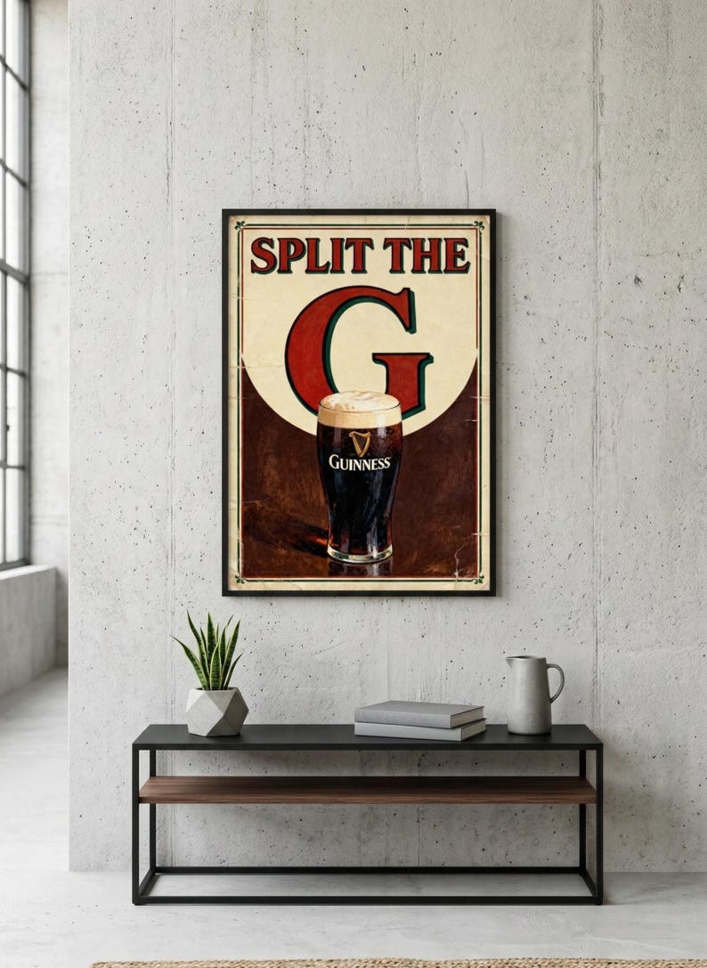 May include: Framed vintage-style Guinness beer advertisement. The poster features a pint of Guinness with a creamy head, and the text "SPLIT THE G". The poster is framed in black and hangs on a concrete wall.