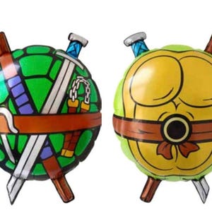 May include: Two round foil balloons, each depicting a Teenage Mutant Ninja Turtle shell. One is green with weapons, the other is yellow. Both have brown accents and are ready for a party.