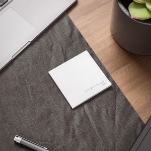 May include: A stack of white square sticky notes with handwritten text in a cursive font. The notes are on a dark grey desk mat next to a laptop and a pen. A potted plant is in the background.
