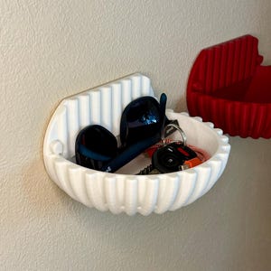 May include: A white, semi-circular wall-mounted storage bowl with a vertical ribbed texture. The bowl contains a pair of black sunglasses and a black car key fob with a red and white lanyard. A second identical red bowl is visible to the right.