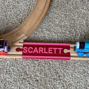May include: A wooden toy train track set featuring a bright pink connector piece with the name SCARLETT embossed in capital letters. The track connects a purple toy locomotive on the left and a blue toy locomotive on the right side.