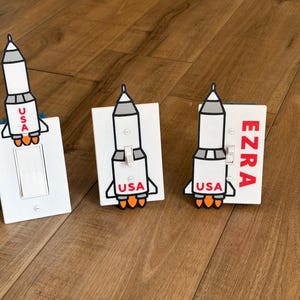 May include: Three white light switch covers with rocket ship cutouts. The rockets are white with gray and black accents, and orange flames. The word "USA" is on each rocket. One cover has the name "EZRA" in red.