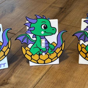 May include: Three decorative light switch covers featuring a cartoon dragon design. The dragons are green with purple accents, sitting in a yellow and brown eggshell. One cover has the name "SCARLETT" on it.