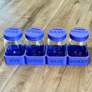 Puede incluir: Cuatro frascos de vidrio transparente con tapas y bases moradas, etiquetados como "GIVE", "SPEND", "SAVE" e "INVEST". Los frascos están dispuestos en fila sobre una superficie de madera. Las tapas tienen ranuras para insertar dinero.