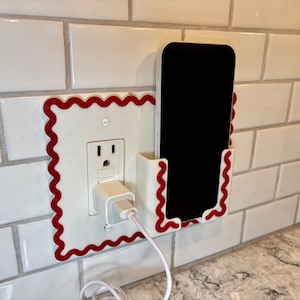 May include: A white wall plate cover with a red wavy border design featuring an integrated vertical phone holder. The plate is mounted over a standard electrical outlet with a white power adapter and charging cable plugged into the socket.