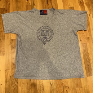 Puede incluir: Camiseta gris jaspeado con un emblema circular azul marino con la inscripción "UNIVERSITY OF OXFORD". La camiseta tiene mangas cortas y cuello redondo. Una etiqueta es visible en el cuello.