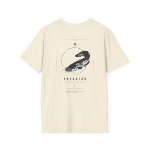 Op de afbeelding: Een crèmekleurig T-shirt met korte mouwen en een zwarte afbeelding van een meerval in een cirkel. De tekst luidt PREDATOR, MASTER OF THE DEEP, SILENT, STRONG, RELENTLESS en FRESHWATER DOMINATOR, samen met kleine geometrische symbolen en lijnen.
