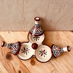 Op de afbeelding: Een decoratieve keramische serveerschaal met een uniek ontwerp. De schaal heeft drie cirkelvormige compartimenten en drie conische, dekselachtige elementen. De schaal is voornamelijk crèmekleurig met rode, zwarte en groene geometrische patronen.