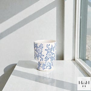 Puede incluir: Una maceta de cer&aacute;mica blanca con un estampado floral azul. La maceta tiene una base de pedestal y una parte superior cil&iacute;ndrica. El dise&ntilde;o floral incluye flores y hojas estilizadas. La maceta est&aacute; sobre una superficie blanca.