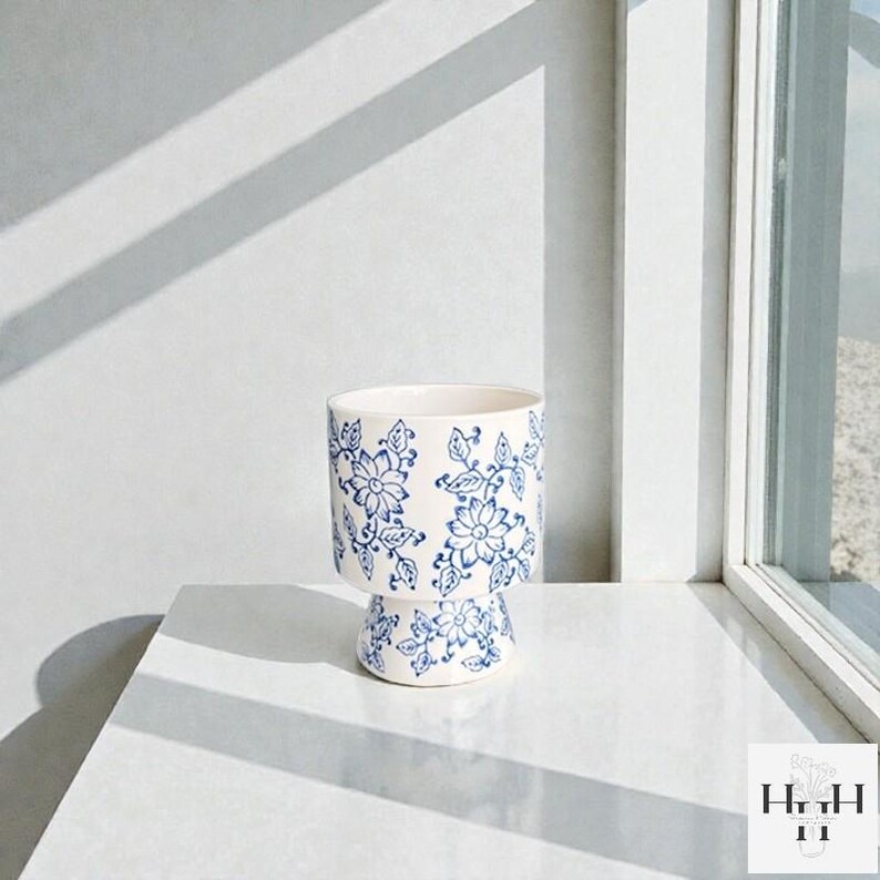Puede incluir: Una maceta de cer&aacute;mica blanca con un estampado floral azul. La maceta tiene una base de pedestal y una parte superior cil&iacute;ndrica. El dise&ntilde;o floral incluye flores y hojas estilizadas. La maceta est&aacute; sobre una superficie blanca.