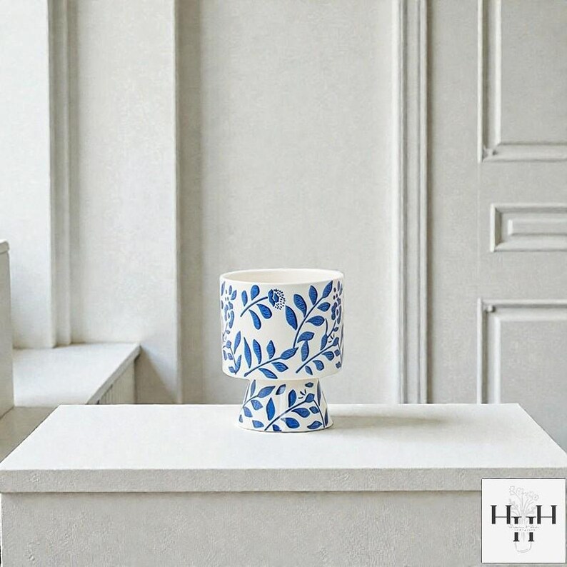 Puede incluir: Una maceta de cer&aacute;mica blanca con un dise&ntilde;o floral azul. La maceta tiene una parte superior cil&iacute;ndrica y una base de pedestal. El patr&oacute;n azul presenta hojas y flores. La maceta est&aacute; sobre una superficie blanca.