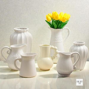 May include: A selection of white ceramic vases and jugs in diverse shapes and sizes. One jug holds a bunch of vibrant yellow tulips. The vases and jugs feature varied textures and handle designs, creating a unified yet diverse arrangement.