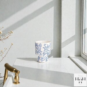 Puede incluir: Maceta de cer&aacute;mica blanca con un dise&ntilde;o floral azul. La maceta tiene una base de pedestal y una parte superior cil&iacute;ndrica. El dise&ntilde;o presenta flores y hojas azules estilizadas. Aproximadamente 15 cm de altura. Para interior o exterior.