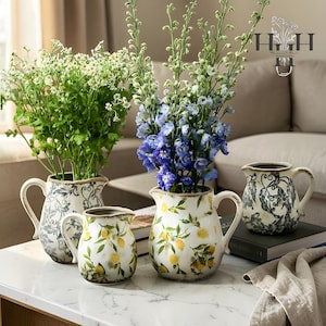 May include: Four ceramic jugs with floral arrangements. Two jugs have blue and white patterns, and two have a lemon design. The jugs are filled with greenery and blue flowers, set on a white table. The jugs have handles.