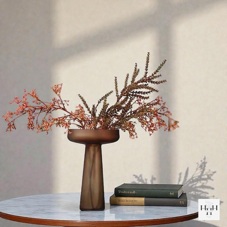 May include: A brown glass vase with a wide, shallow top and a slender stem, holding dried floral branches with reddish-orange and green hues. The vase sits on a marble-topped table, next to two books. The overall aesthetic is minimalist and modern.