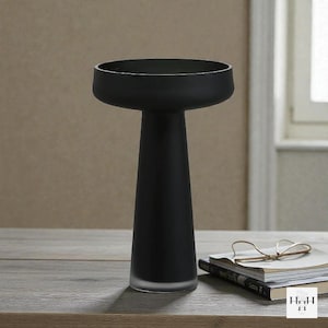 May include: A matte black decorative vase with a wide, shallow bowl and a tall, conical stem. The vase is made of glass and has a frosted base. It sits on a wooden surface with books and glasses.