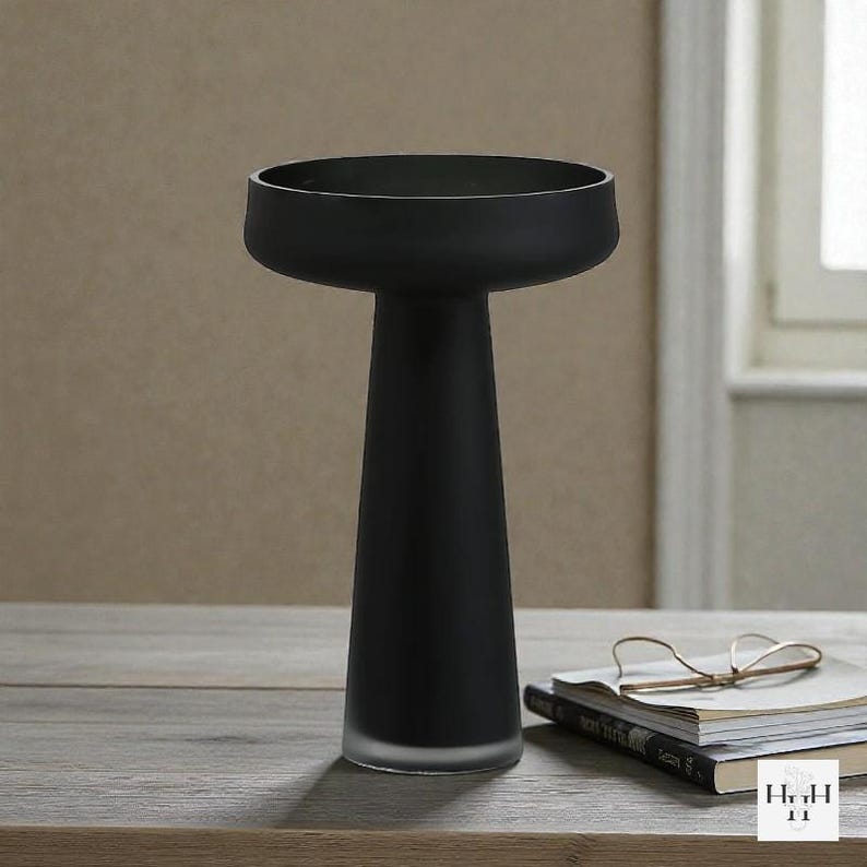 May include: A matte black decorative vase with a wide, shallow bowl and a tall, conical stem. The vase is made of glass and has a frosted base. It sits on a wooden surface with books and glasses.