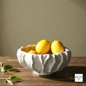 May include: A white ceramic bowl with a textured, wavy exterior, filled with bright yellow lemons. The bowl sits on a wooden surface, with green leaves in the foreground. The bowl's interior is a contrasting yellow.