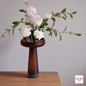 May include: A brown, frosted glass vase with a wide, shallow top and a tall, slender base. The vase holds white flowers and green foliage. The arrangement sits on a wooden surface.